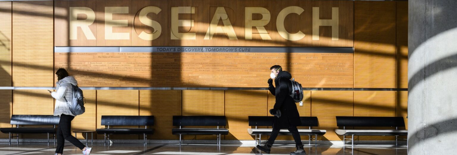 Sun-drenched wood wall with the word Research on it in the background with two people walking in the foreground