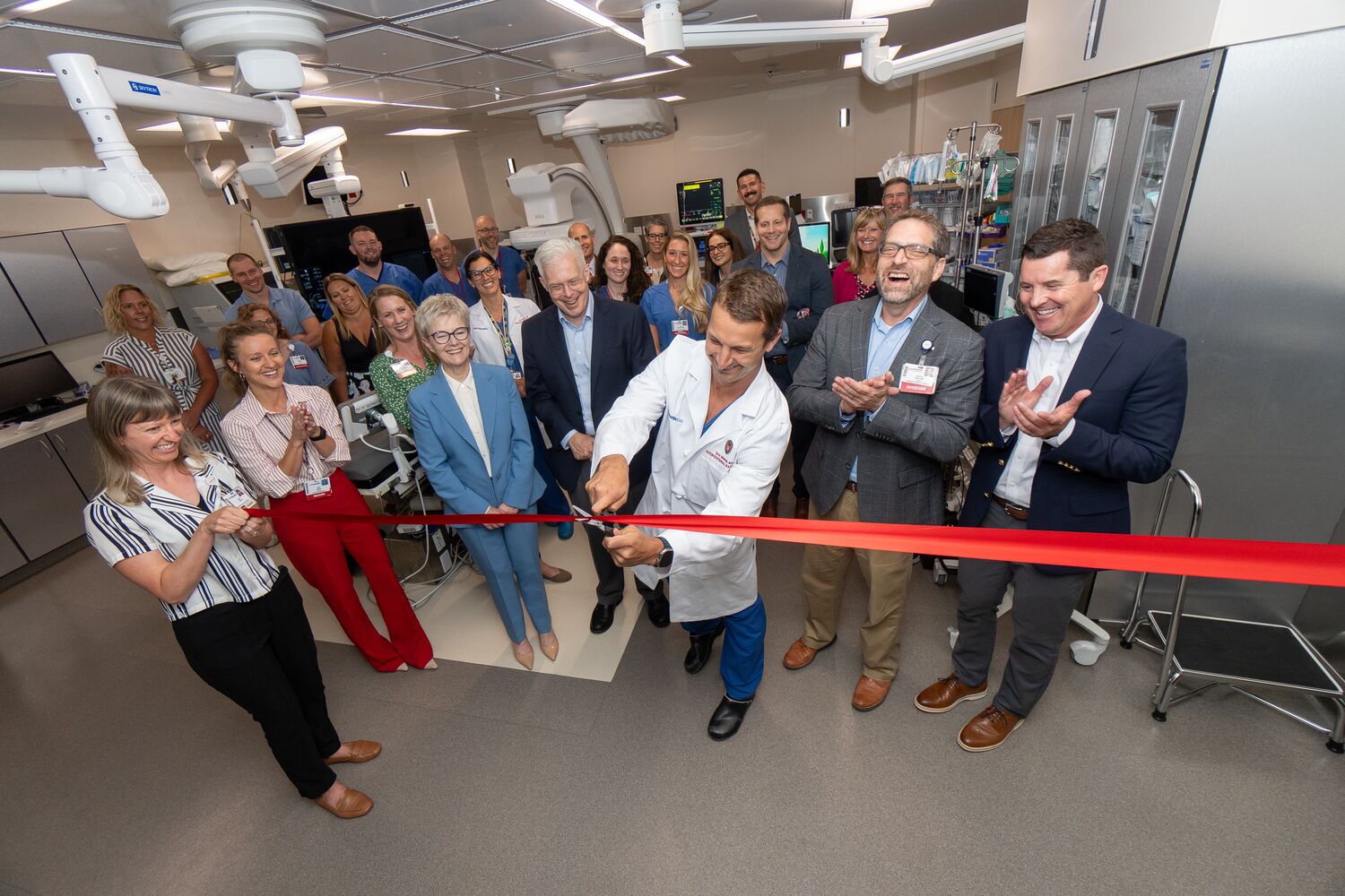 A doctor in a white coat cuts a ribbon to celebrate the opening of a new pediatric interventional radiology suite. The group of people are standing in the medical suite. 
