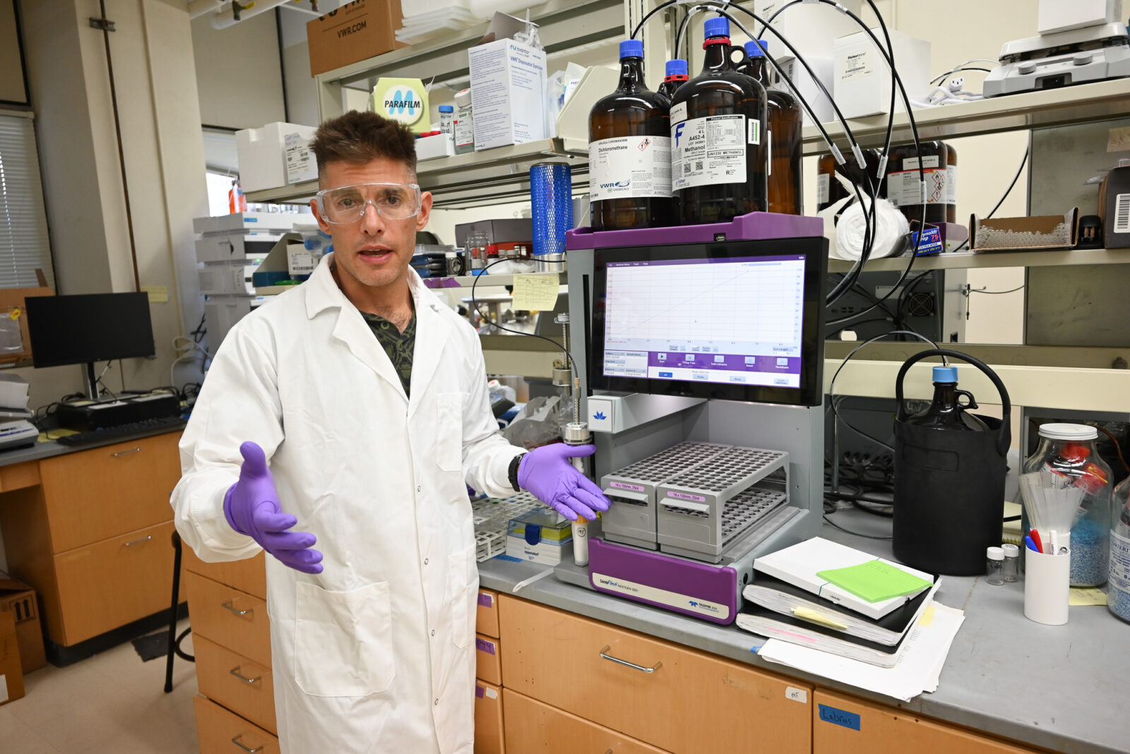 A scientist talks about lab equipment while in front of a monitor in a lab. He is wearing a white coat and safety goggles.