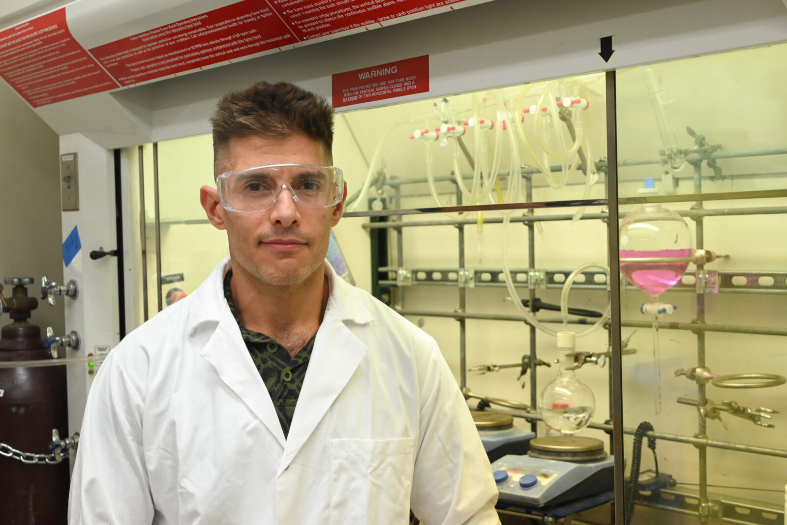 A scientist poses for a photo in his lab. He is wearing a white coat and safety goggles.