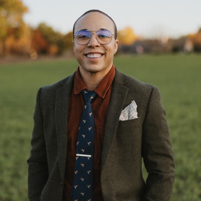 A man in a suit jacket, tie, and glasses smiles for the camera. He is standing outside on a green lawn.